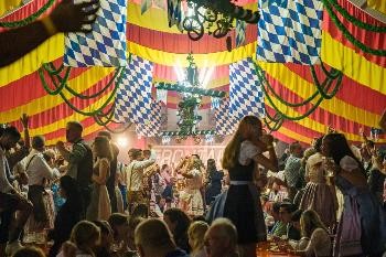 Blick in ein bunt dekoriertes Festzelt, gefüllt mit Menschen, die teilweise auf den Baänken stehen und fröhlich feiern